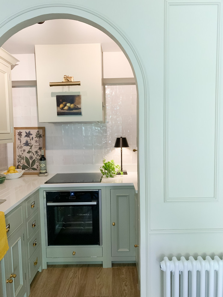Photo of a small classic u-shaped kitchen/diner in Kent with a built-in sink, raised-panel cabinets, green cabinets, quartz worktops, white splashback, ceramic splashback, black appliances, medium hardwood flooring, white worktops and feature lighting.
