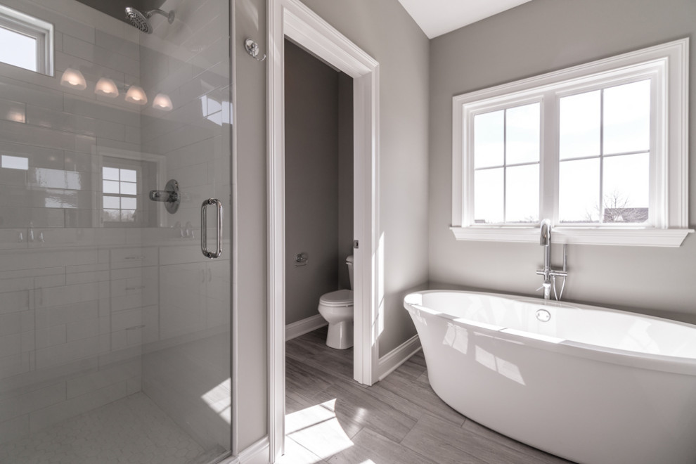 This is an example of a transitional master bathroom in Louisville with recessed-panel cabinets, grey cabinets, a freestanding tub, an alcove shower, a one-piece toilet, white tile, subway tile, grey walls, wood-look tile, a drop-in sink, solid surface benchtops, grey floor, a hinged shower door, white benchtops, an enclosed toilet, a double vanity and a built-in vanity.