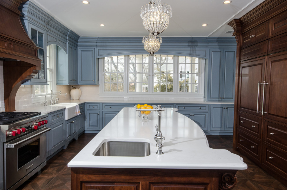 Photo of a large traditional u-shaped eat-in kitchen in New York with a farmhouse sink, recessed-panel cabinets, blue cabinets, solid surface benchtops, white splashback, ceramic splashback, dark hardwood floors, with island, panelled appliances and brown floor.