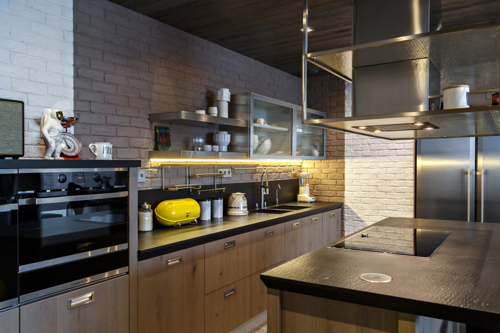 Photo of a mid-sized industrial galley kitchen in Moscow with a double-bowl sink, flat-panel cabinets, medium wood cabinets, grey splashback, brick splashback, stainless steel appliances, with island, black benchtop and wood.
