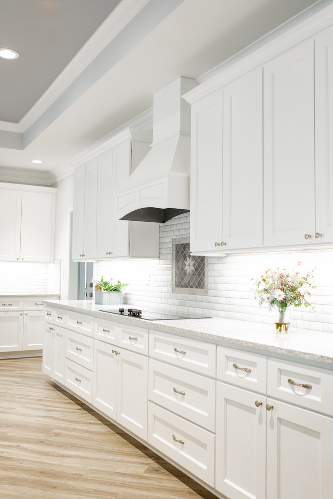 This is an example of an expansive transitional l-shaped eat-in kitchen in Charlotte with a triple-bowl sink, shaker cabinets, white cabinets, quartz benchtops, white splashback, marble splashback, stainless steel appliances, light hardwood floors, with island, grey floor and multi-coloured benchtop.