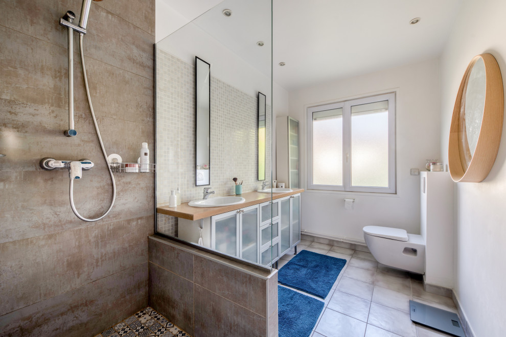 Photo of a contemporary bathroom in Paris with glass-front cabinets, white cabinets, a curbless shower, a wall-mount toilet, beige tile, white tile, brown tile, white walls, porcelain floors, a vessel sink, wood benchtops, grey floor, an open shower and brown benchtops.