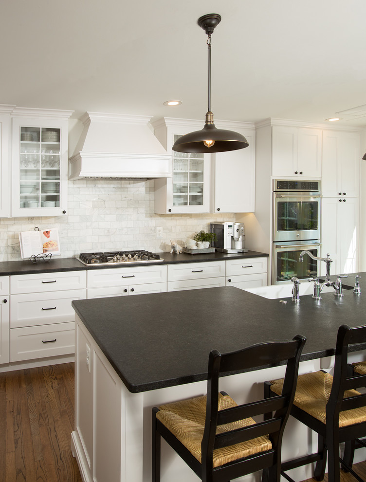 This is an example of a mid-sized traditional l-shaped eat-in kitchen in DC Metro with a farmhouse sink, flat-panel cabinets, white cabinets, granite benchtops, grey splashback, marble splashback, stainless steel appliances, dark hardwood floors, with island and brown floor.