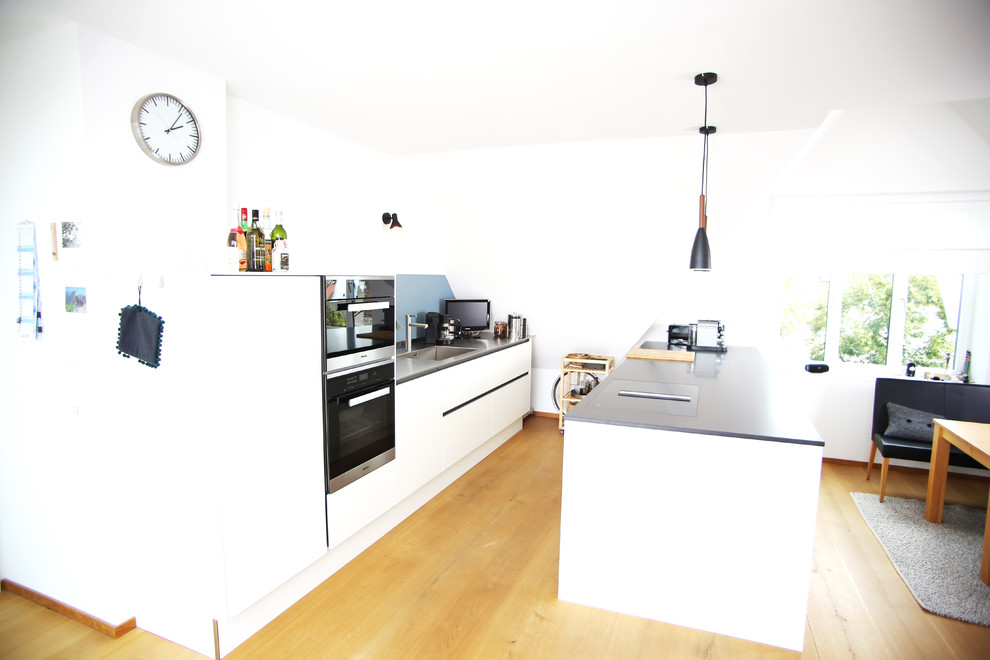 This is an example of a mid-sized modern galley eat-in kitchen in Munich with a single-bowl sink, flat-panel cabinets, white cabinets, stainless steel benchtops, stainless steel appliances and with island.