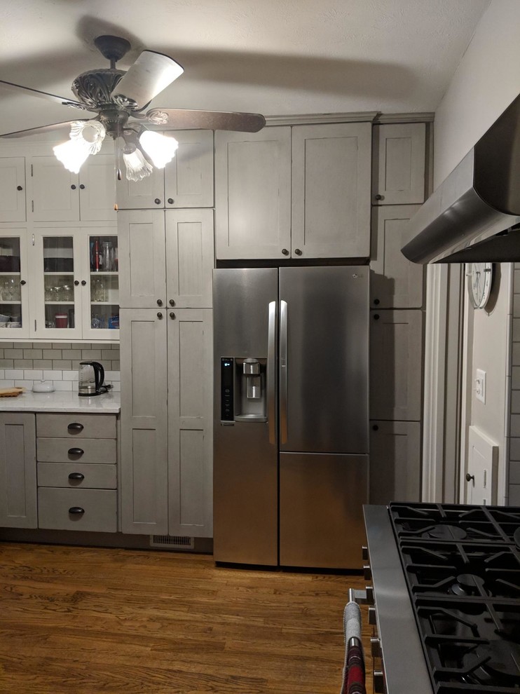 Small traditional l-shaped eat-in kitchen in Omaha with a farmhouse sink, shaker cabinets, grey cabinets, quartz benchtops, white splashback, porcelain splashback, stainless steel appliances, medium hardwood floors, brown floor and white benchtop.