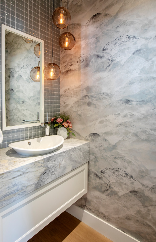 Small arts and crafts powder room in Vancouver with shaker cabinets, white cabinets, a wall-mount toilet, blue tile, porcelain tile, multi-coloured walls, medium hardwood floors, a vessel sink, marble benchtops, brown floor and blue benchtops.