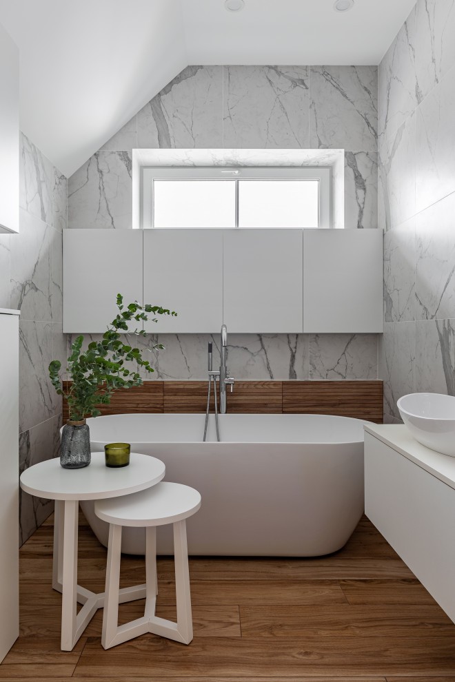 Inspiration pour une salle de bain design avec un placard à porte plane, des portes de placard blanches, une baignoire indépendante, un carrelage gris, un sol en bois brun, une vasque, un sol marron, un plan de toilette blanc et meuble-lavabo suspendu.