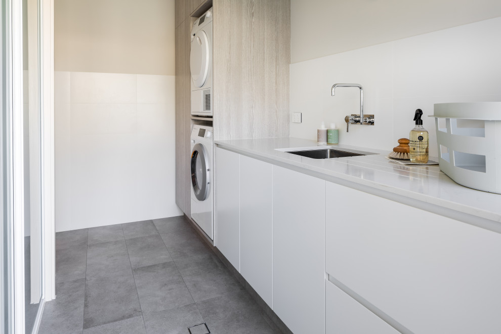 Photo of a mid-sized contemporary single-wall utility room in Perth with an undermount sink, flat-panel cabinets, quartz benchtops, white splashback, porcelain splashback, porcelain floors, a stacked washer and dryer, grey floor and white benchtop.