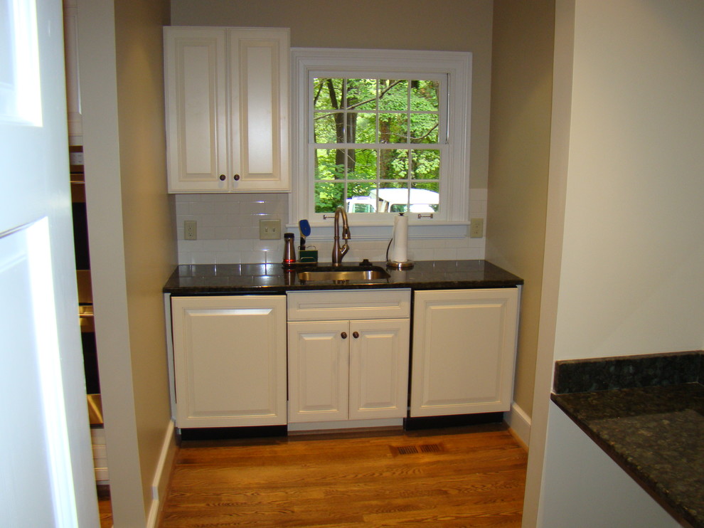 Home design - small transitional single-wall medium tone wood floor home design idea in Charlotte with an undermount sink, raised-panel cabinets, white cabinets, granite countertops, white backsplash and subway tile backsplash