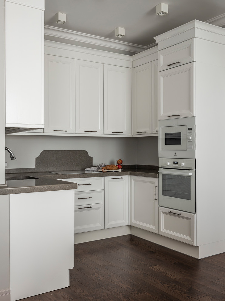 Design ideas for a small transitional u-shaped kitchen in Moscow with an undermount sink, recessed-panel cabinets, white cabinets, grey splashback, white appliances, dark hardwood floors, no island, brown floor and grey benchtop.