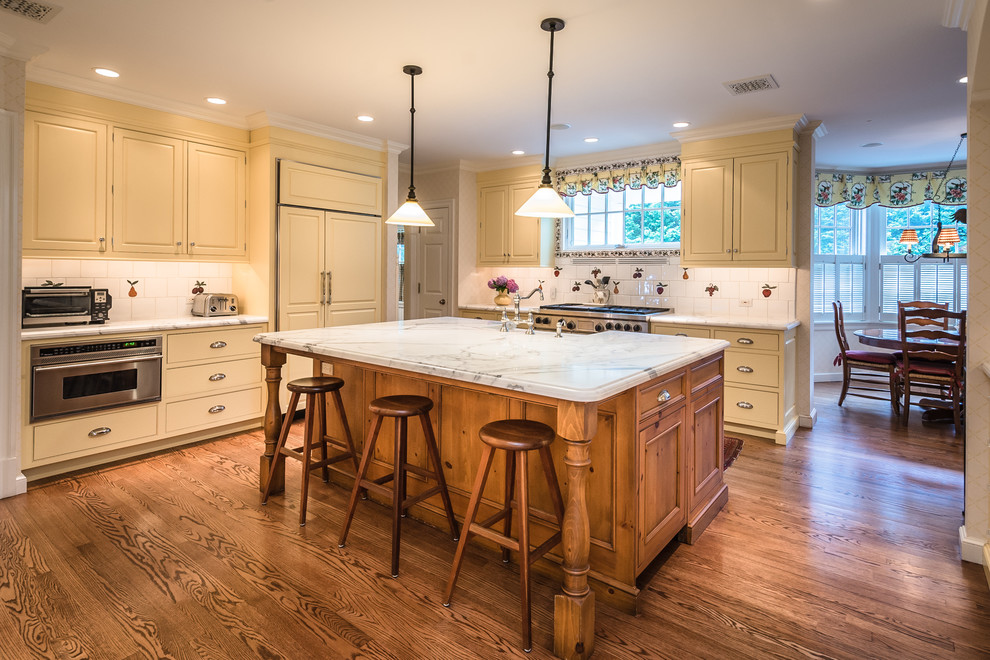 This is an example of a large traditional l-shaped eat-in kitchen in New York with a drop-in sink, beaded inset cabinets, medium wood cabinets, marble benchtops, multi-coloured splashback, stone slab splashback, stainless steel appliances, medium hardwood floors, with island and brown floor.