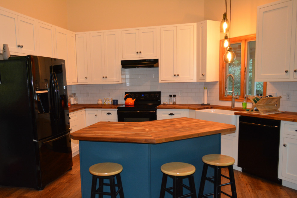 Example of a small transitional u-shaped medium tone wood floor and brown floor eat-in kitchen design with a farmhouse sink, recessed-panel cabinets, white cabinets, wood countertops, white backsplash, ceramic backsplash, black appliances, an island and brown countertops