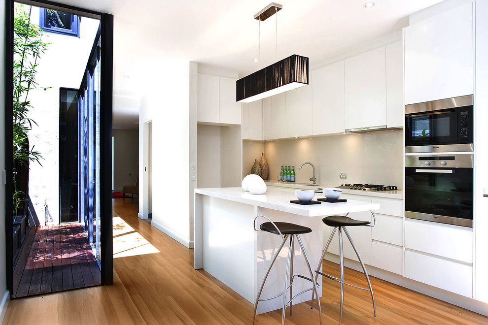 Design ideas for a small contemporary galley eat-in kitchen in Sydney with flat-panel cabinets, white cabinets, stainless steel appliances, an undermount sink, quartz benchtops, beige splashback, glass sheet splashback, medium hardwood floors and with island.