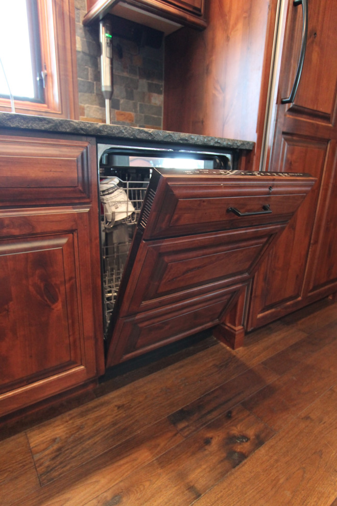 Example of a large mountain style medium tone wood floor and brown floor kitchen design with raised-panel cabinets, medium tone wood cabinets, granite countertops, multicolored backsplash, stone tile backsplash, paneled appliances, an island and black countertops