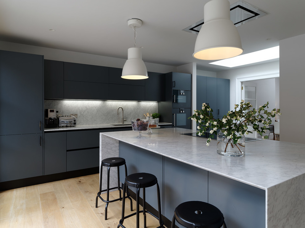 Large contemporary single-wall open plan kitchen in Sussex with flat-panel cabinets, blue cabinets, marble benchtops, grey splashback, marble splashback, with island, grey benchtop, an integrated sink, black appliances, light hardwood floors and brown floor.