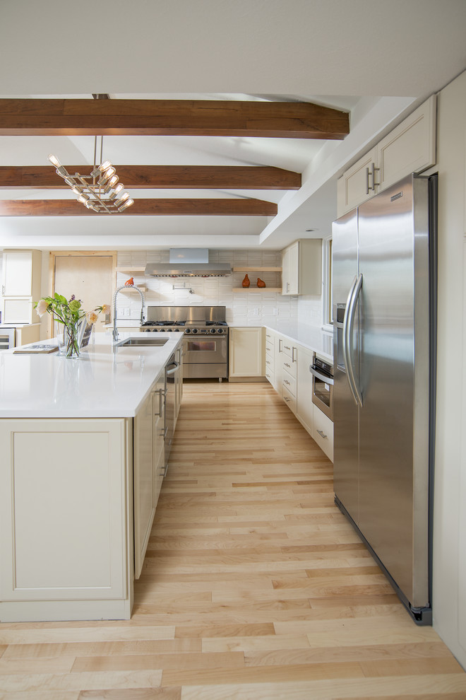 This is an example of a transitional l-shaped eat-in kitchen in Other with an undermount sink, recessed-panel cabinets, white cabinets, solid surface benchtops, white splashback, ceramic splashback, stainless steel appliances, light hardwood floors and with island.