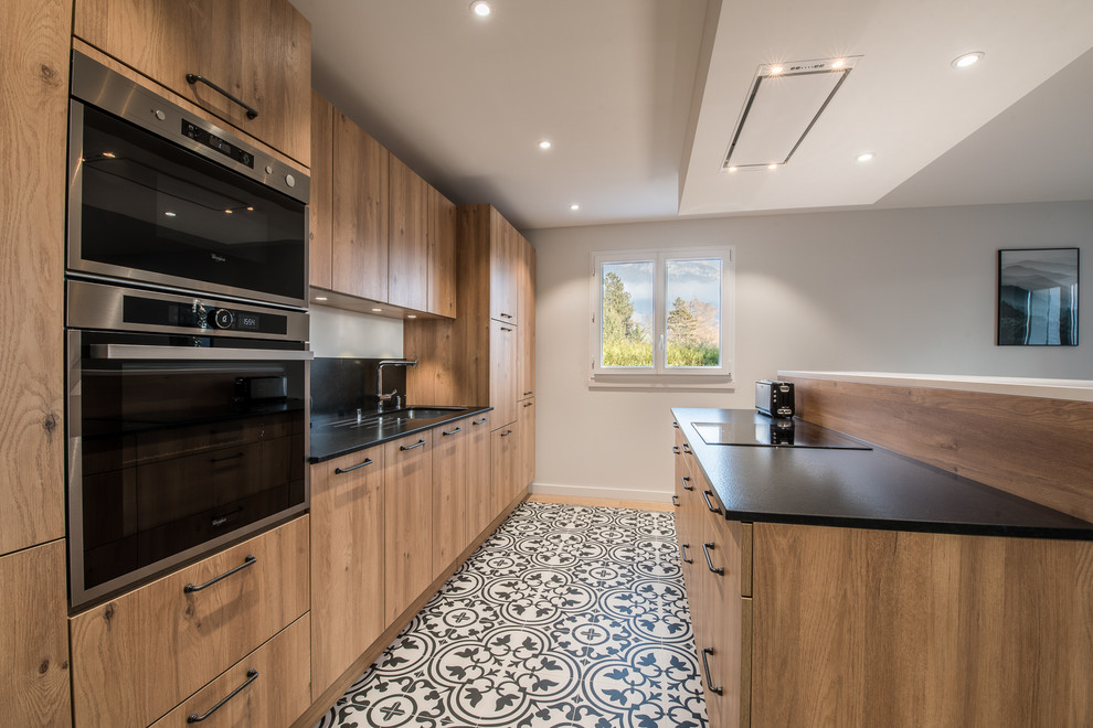 Inspiration for a mid-sized contemporary galley open plan kitchen in Grenoble with an undermount sink, flat-panel cabinets, medium wood cabinets, black appliances, cement tiles, with island and black splashback.