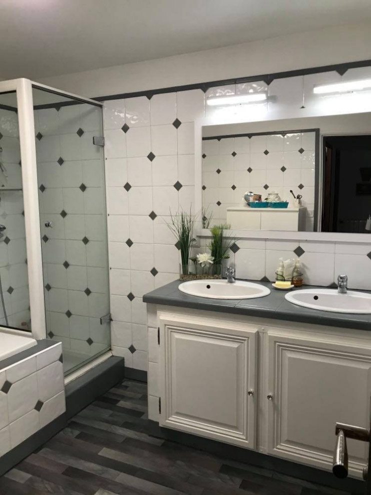 Пример оригинального дизайна: серо-белая ванная комната с белыми фасадами, полновстраиваемой ванной, душевой комнатой, белой плиткой, керамической плиткой, белыми стенами, полом из линолеума, врезной раковиной, столешницей из плитки, серым полом, душем с распашными дверями, серой столешницей, тумбой под две раковины и встроенной тумбой