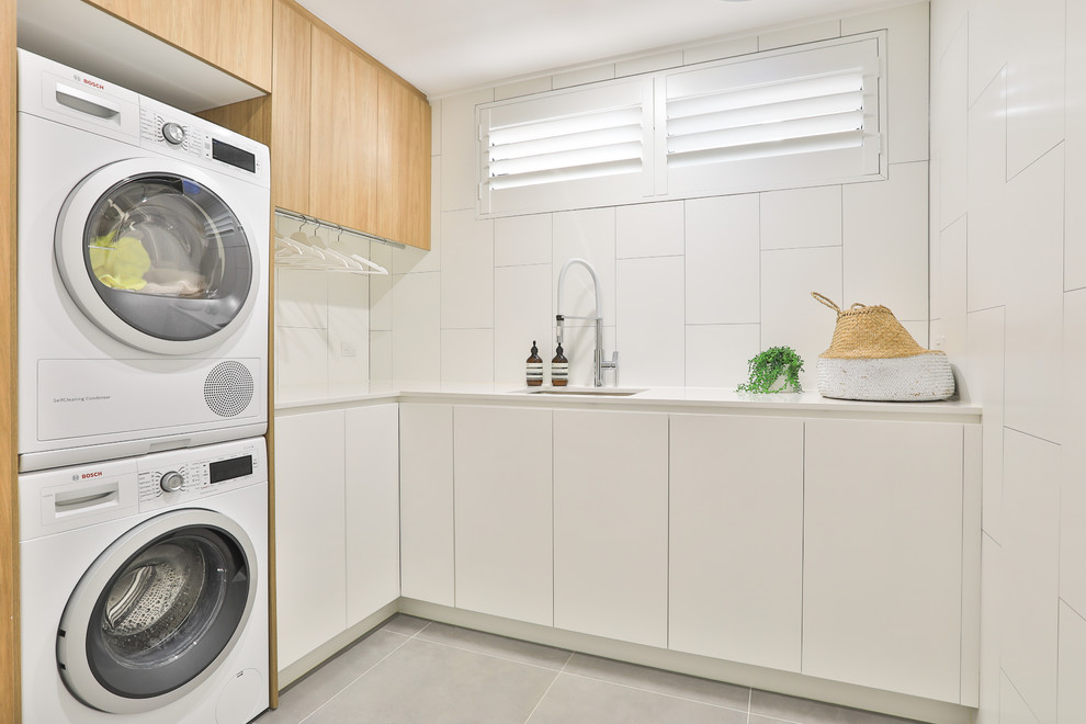 Photo of a contemporary l-shaped dedicated laundry room in Sydney with an undermount sink, flat-panel cabinets, white cabinets, quartz benchtops, white walls, ceramic floors, a stacked washer and dryer, grey floor and white benchtop.