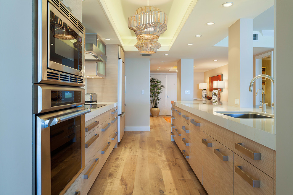 This is an example of a large contemporary galley open plan kitchen in Miami with an undermount sink, flat-panel cabinets, light wood cabinets, solid surface benchtops, white splashback, stainless steel appliances, light hardwood floors, no island, beige floor and white benchtop.