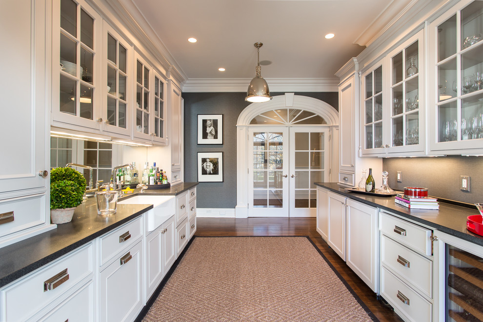 This is an example of a traditional galley wet bar in New York with glass-front cabinets, white cabinets, grey splashback, dark hardwood floors and brown floor.