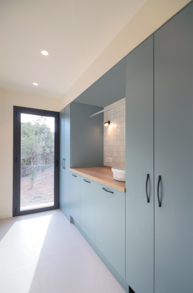 Mid-sized contemporary single-wall dedicated laundry room in Barcelona with a farmhouse sink, flat-panel cabinets, turquoise cabinets, wood benchtops, white splashback, ceramic splashback, white walls, ceramic floors, a concealed washer and dryer, beige floor and brown benchtop.