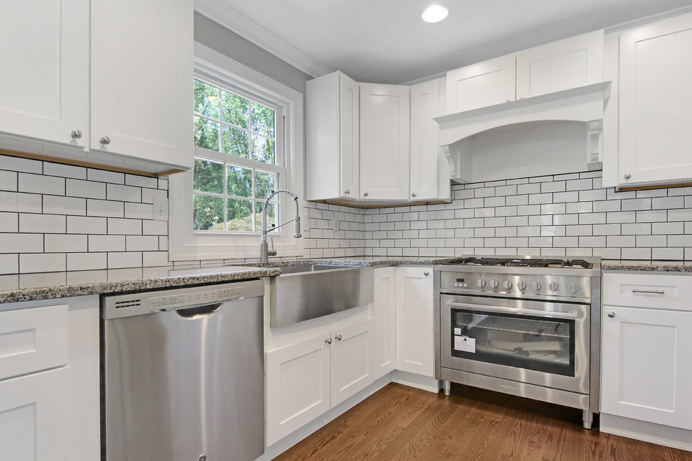 Photo of a large country l-shaped eat-in kitchen in Philadelphia with a farmhouse sink, shaker cabinets, white cabinets, granite benchtops, white splashback, subway tile splashback, stainless steel appliances, medium hardwood floors, with island, brown floor and black benchtop.