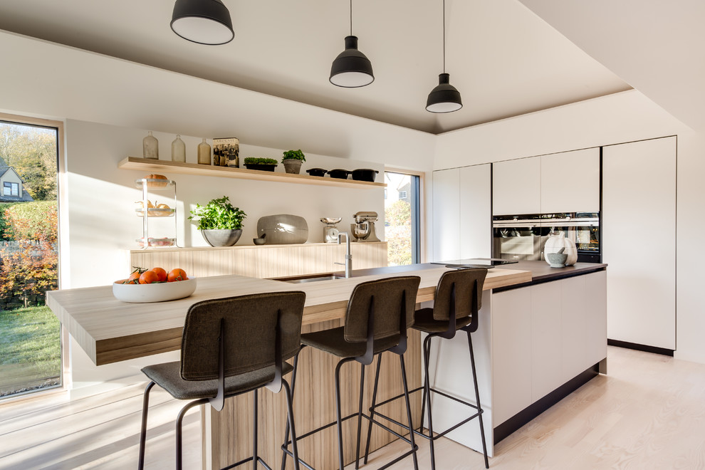 Photo of a mid-sized contemporary single-wall kitchen in Gloucestershire with an undermount sink, flat-panel cabinets, black appliances, light hardwood floors, with island, multi-coloured benchtop and beige floor.