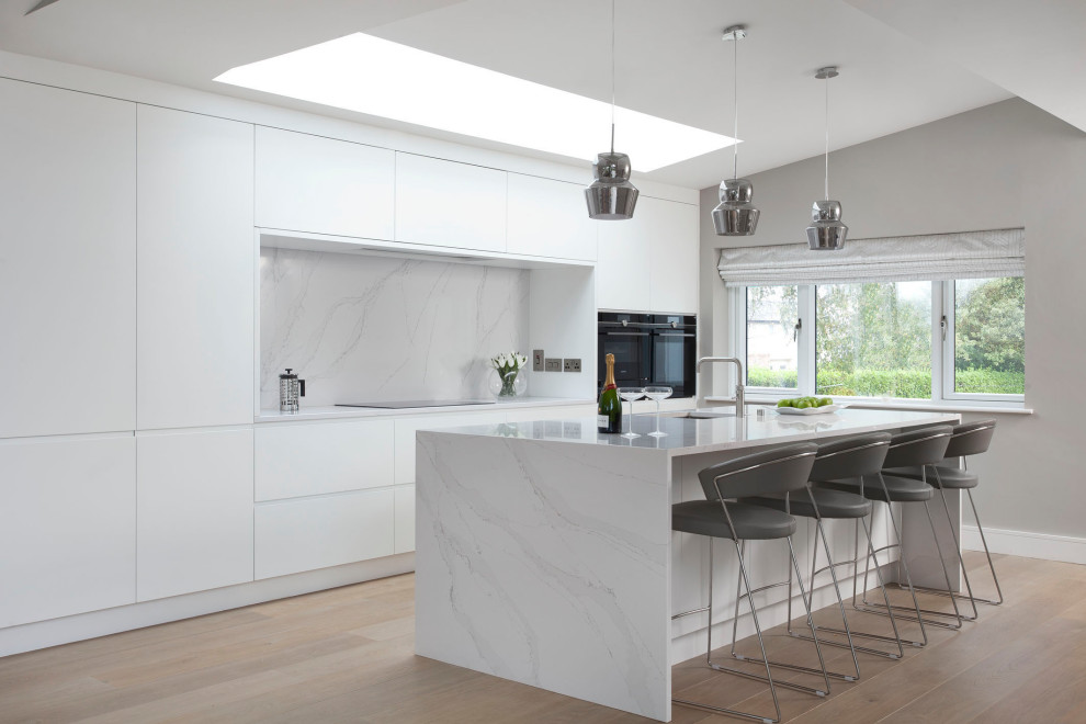 Design ideas for a contemporary galley kitchen in Dublin with an undermount sink, flat-panel cabinets, white cabinets, white splashback, stone slab splashback, black appliances, light hardwood floors, with island, beige floor and white benchtop.