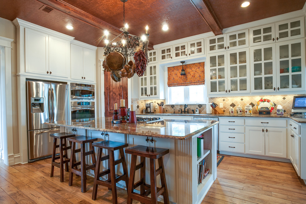 Large country u-shaped open plan kitchen in Dallas with a farmhouse sink, glass-front cabinets, white cabinets, granite benchtops, beige splashback, ceramic splashback, stainless steel appliances, medium hardwood floors and with island.