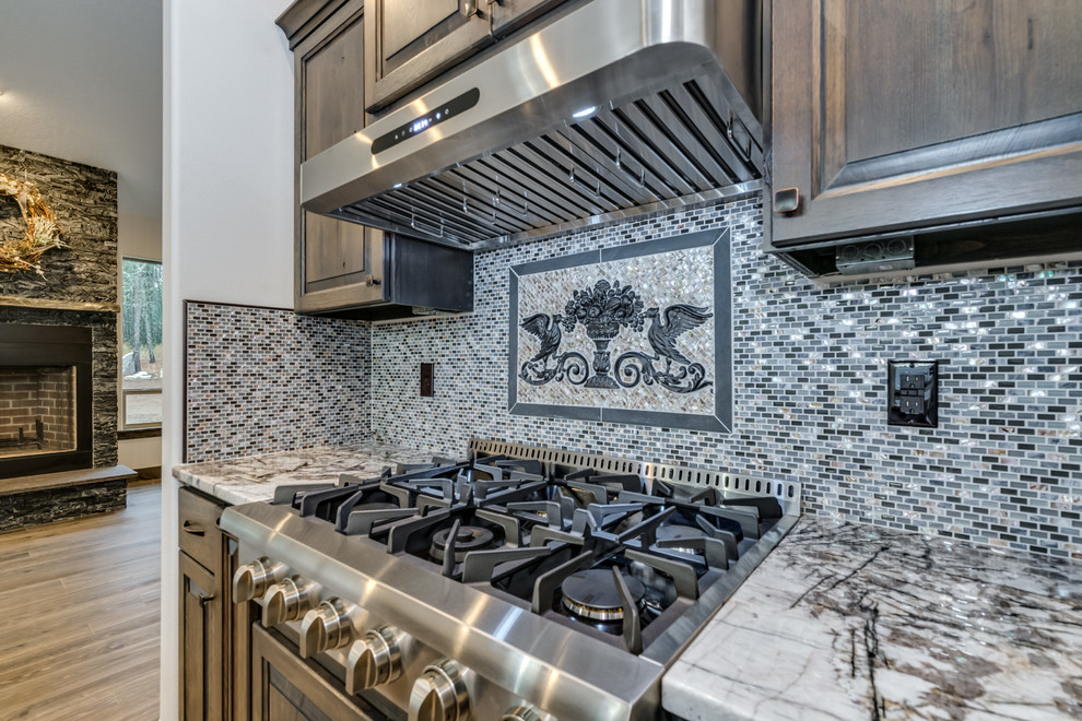 Example of a large mountain style u-shaped laminate floor and brown floor open concept kitchen design with an undermount sink, raised-panel cabinets, medium tone wood cabinets, quartz countertops, gray backsplash, mosaic tile backsplash, stainless steel appliances and an island