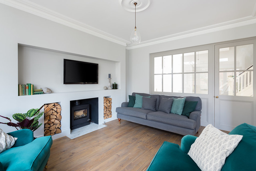 Photo of a transitional living room in London with white walls, medium hardwood floors, a wood stove, a wall-mounted tv and brown floor.