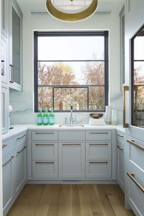 Light gray Shaker kitchen cabinets showing classic five-piece doors—two stiles, two rails, and a recessed floating center panel—with brass pulls beneath a large window