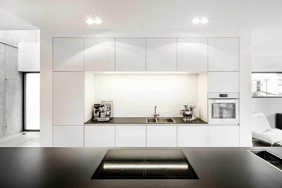 Modern open plan kitchen in Cologne with a double-bowl sink, flat-panel cabinets, white splashback, with island, white appliances and concrete floors.