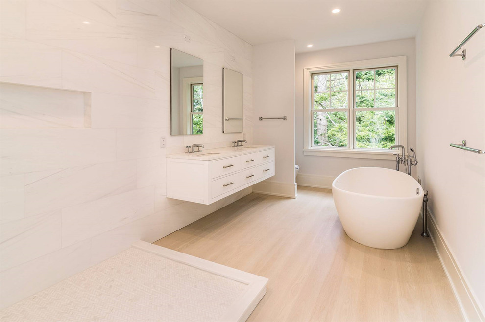Expansive contemporary master bathroom in New York with furniture-like cabinets, white cabinets, a freestanding tub, an open shower, a two-piece toilet, white tile, white walls, light hardwood floors, an undermount sink, beige floor and an open shower.