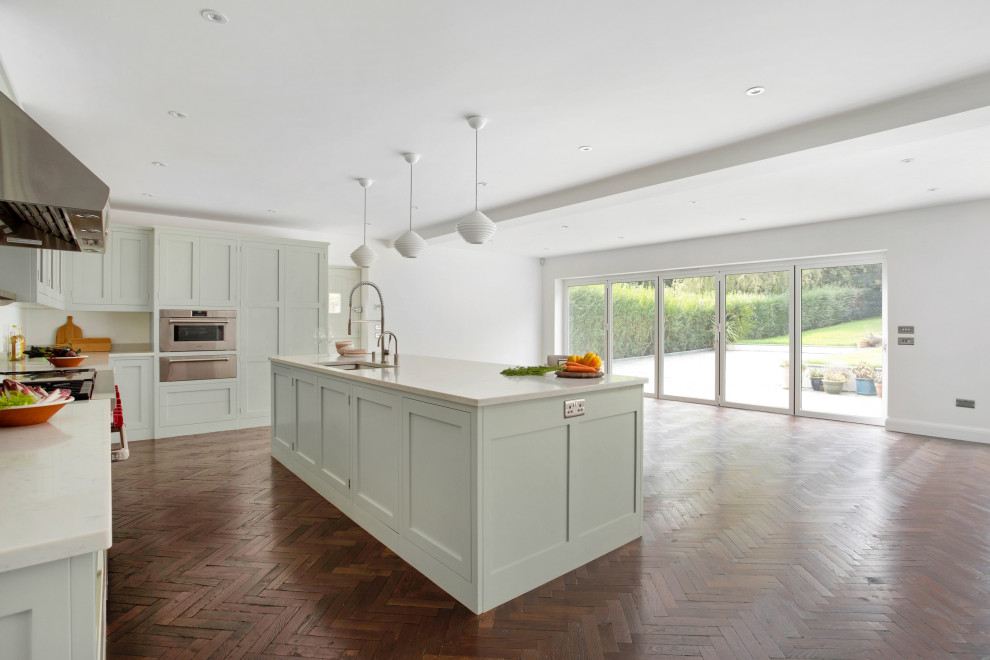 Inspiration for a large classic u-shaped open plan kitchen in London with a submerged sink, shaker cabinets, green cabinets, quartz worktops, stainless steel appliances, medium hardwood flooring, an island, brown floors and white worktops.