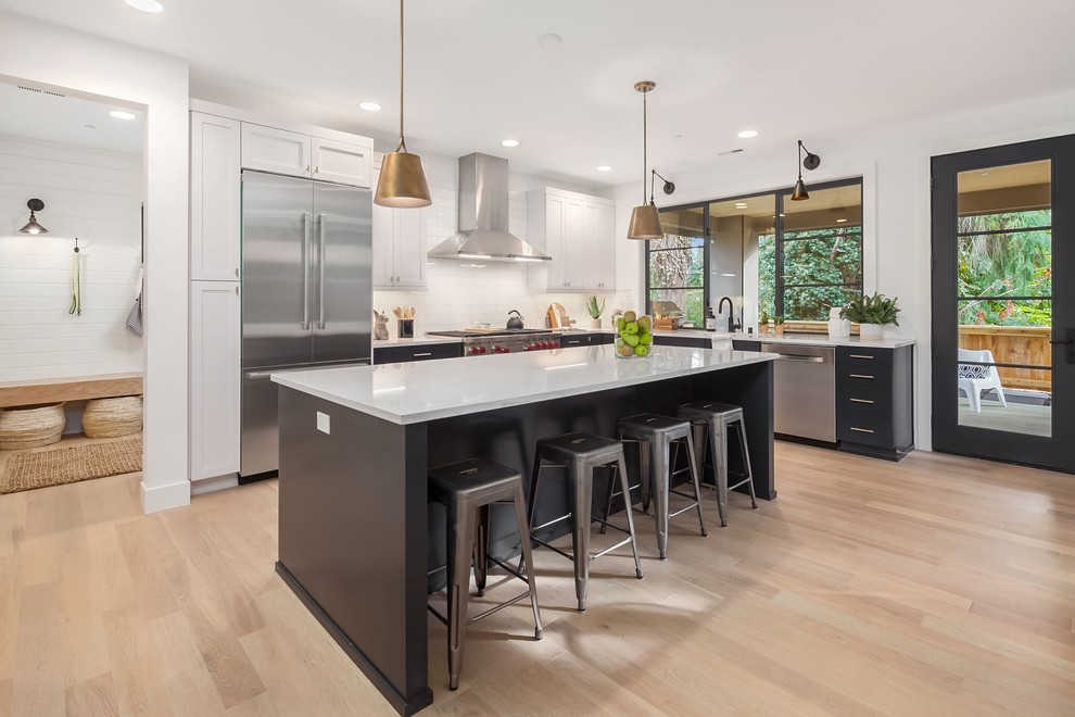 Design ideas for a large transitional l-shaped eat-in kitchen in Seattle with shaker cabinets, white splashback, stainless steel appliances, light hardwood floors, with island, white benchtop, an undermount sink, granite benchtops, porcelain splashback and brown floor.