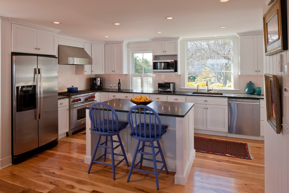 Photo of a large beach style l-shaped eat-in kitchen in Boston with stainless steel appliances, an undermount sink, shaker cabinets, white cabinets, white splashback, subway tile splashback, medium hardwood floors, with island, quartz benchtops and brown floor.