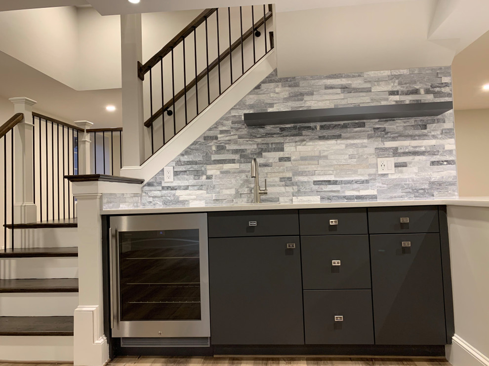 This is an example of a mid-sized transitional l-shaped wet bar in Boston with an undermount sink, flat-panel cabinets, grey cabinets, solid surface benchtops, grey splashback, stone tile splashback, medium hardwood floors, brown floor and white benchtop.