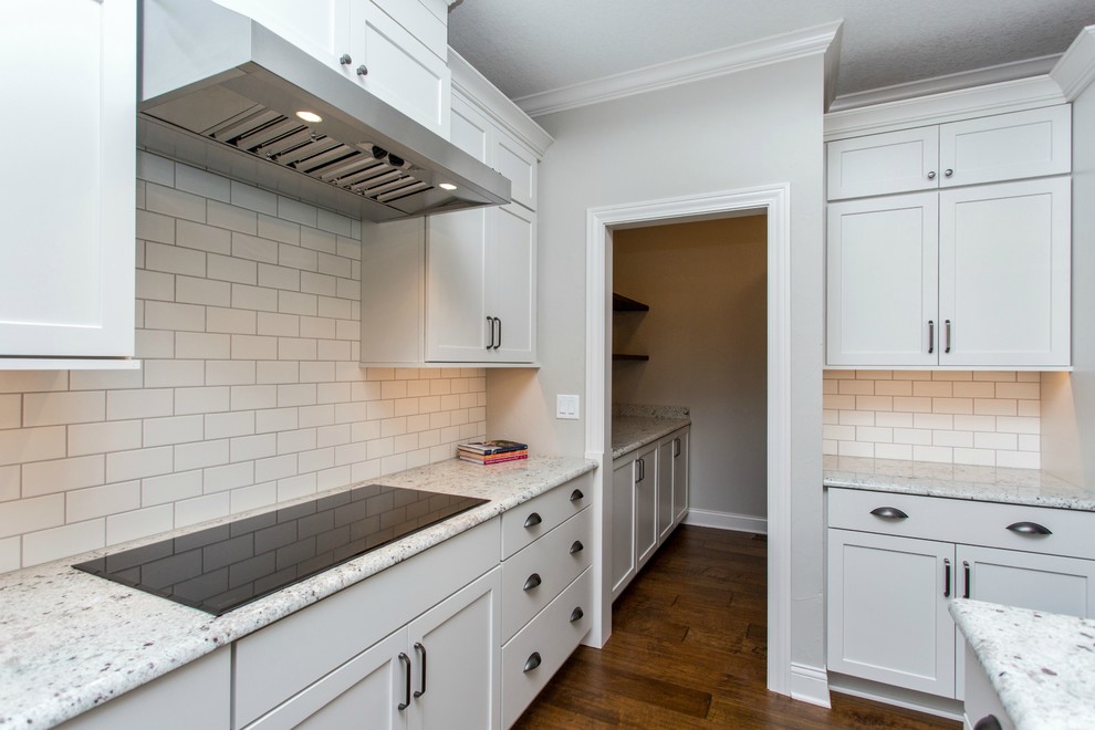 Example of a mid-sized transitional single-wall medium tone wood floor and beige floor open concept kitchen design with a farmhouse sink, recessed-panel cabinets, white cabinets, granite countertops, white backsplash, ceramic backsplash, stainless steel appliances and an island