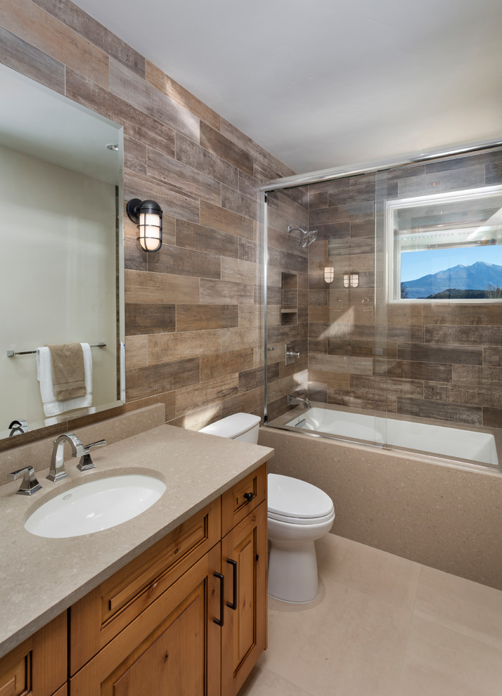 Mid-sized traditional 3/4 bathroom in Denver with raised-panel cabinets, medium wood cabinets, an alcove tub, a shower/bathtub combo, brown tile, porcelain tile, brown walls, limestone floors, an undermount sink, limestone benchtops, beige floor and a sliding shower screen.