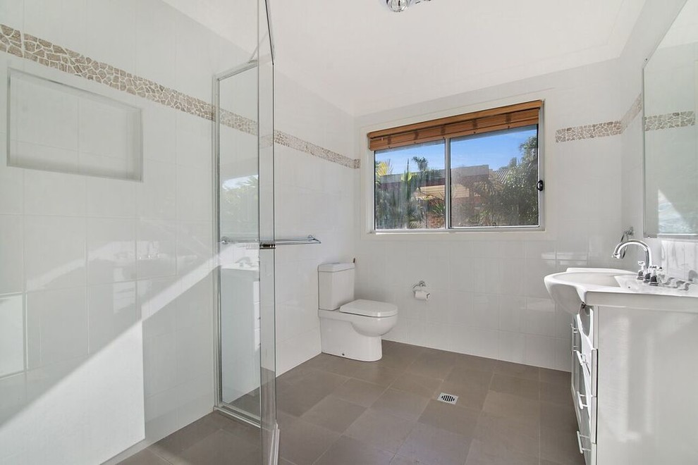 Photo of a mid-sized beach style kids bathroom in Sydney with glass-front cabinets, white cabinets, an alcove shower, a one-piece toilet, gray tile, ceramic tile, white walls, ceramic floors, a wall-mount sink and solid surface benchtops.