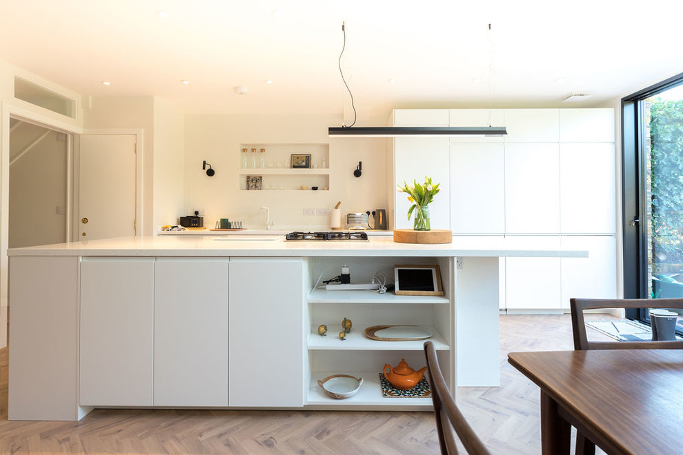 This is an example of a mid-sized contemporary single-wall open plan kitchen in London with flat-panel cabinets, white cabinets, solid surface benchtops, light hardwood floors, with island, brown floor and white benchtop.