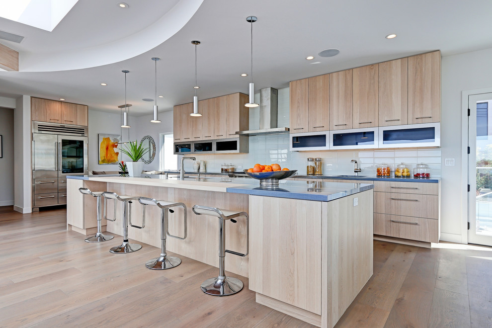 This is an example of a large contemporary open plan kitchen in Los Angeles with a drop-in sink, flat-panel cabinets, light wood cabinets, quartzite benchtops, white splashback, glass tile splashback, stainless steel appliances, light hardwood floors and with island.
