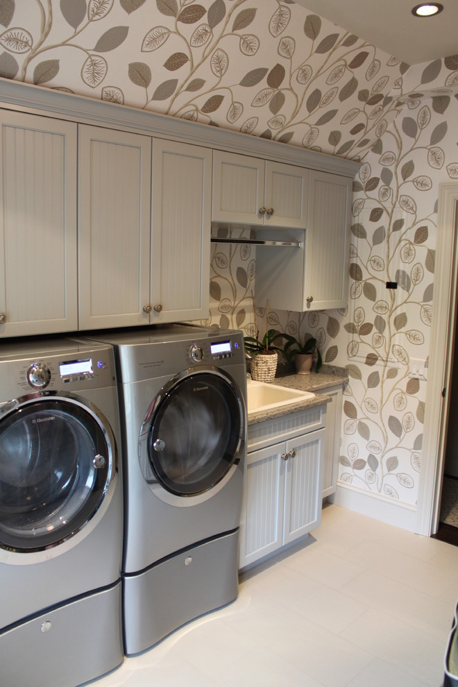 This is an example of a traditional galley dedicated laundry room in Boston with a drop-in sink, recessed-panel cabinets, grey cabinets, beige walls, porcelain floors and a side-by-side washer and dryer.