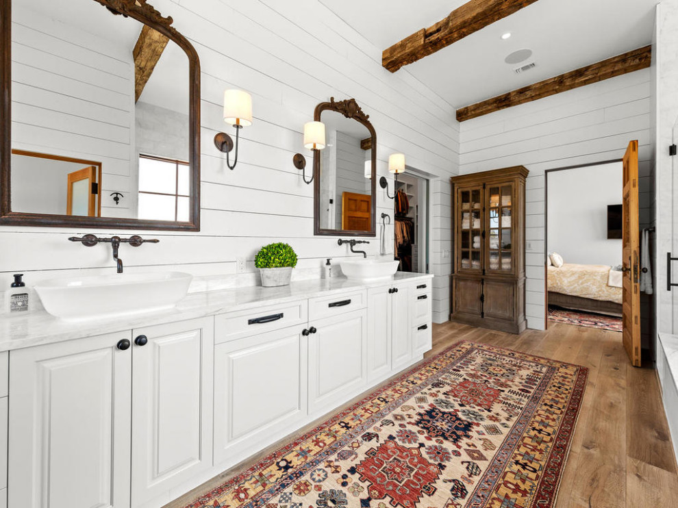 Design ideas for a country bathroom in Austin with raised-panel cabinets, white cabinets, a drop-in tub, gray tile, marble, white walls, medium hardwood floors, a vessel sink, marble benchtops, beige floor, a hinged shower door, grey benchtops, a double vanity, exposed beam and planked wall panelling.