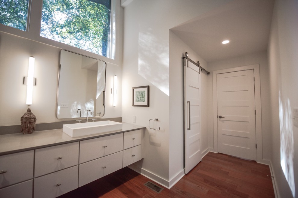 Inspiration for a large transitional master bathroom in Atlanta with flat-panel cabinets, white cabinets, a freestanding tub, a shower/bathtub combo, a one-piece toilet, gray tile, travertine, white walls, dark hardwood floors, a vessel sink, quartzite benchtops, brown floor and a hinged shower door.