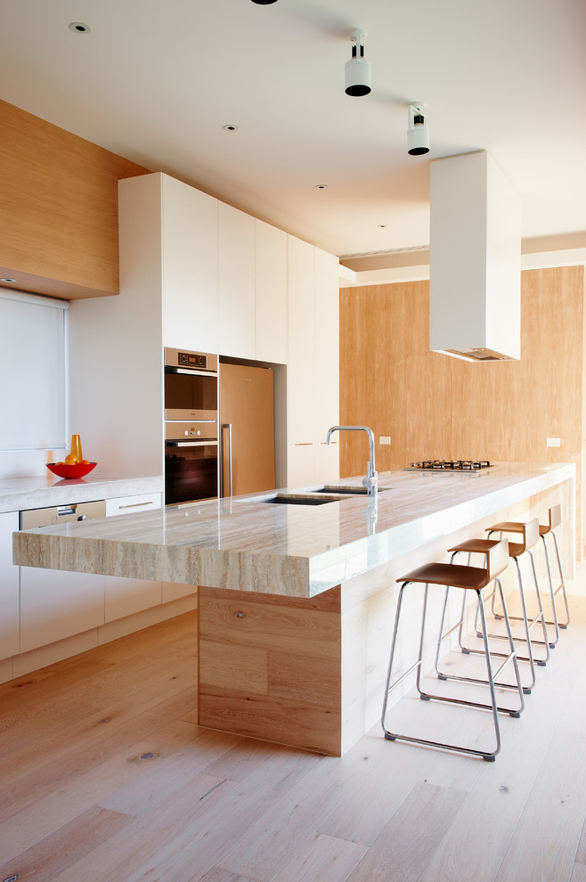 This is an example of a large modern galley kitchen in Melbourne with a double-bowl sink, white cabinets, light hardwood floors, with island, flat-panel cabinets and panelled appliances.