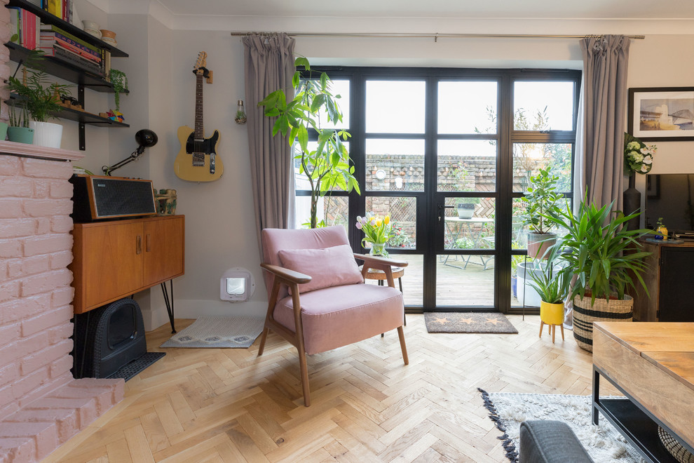 This is an example of a mid-sized eclectic enclosed living room in London with pink walls, medium hardwood floors, a standard fireplace, a brick fireplace surround, a freestanding tv, brown floor and brick walls.