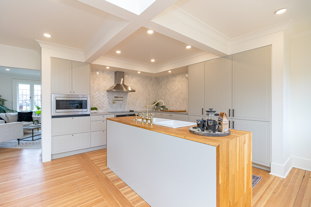 Example of a mid-sized transitional l-shaped medium tone wood floor, brown floor and exposed beam eat-in kitchen design with a drop-in sink, raised-panel cabinets, gray cabinets, wood countertops, beige backsplash, ceramic backsplash, stainless steel appliances, an island and brown countertops